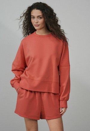 Frau mit lockigem Haar, die einen lockeren roten Pullover und passende rote Shorts trägt, vor einem einfarbigen hellgrauen Hintergrund steht.