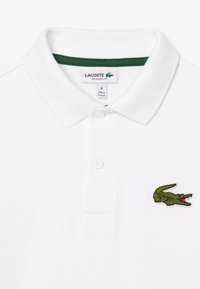 Polo shirt blanc en tissu texturé, col vert, deux boutons et logo de crocodile vert brodé sur la poitrine gauche.