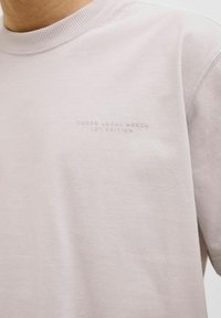 Šviesiai rožinė trumpomis rankovėmis marškinėliai, pagaminti iš švelnios medvilnės, su šone banguotu kaklu ir reljefiniu užrašu "CORE® LOCAL MERCH LDT. EDITION."