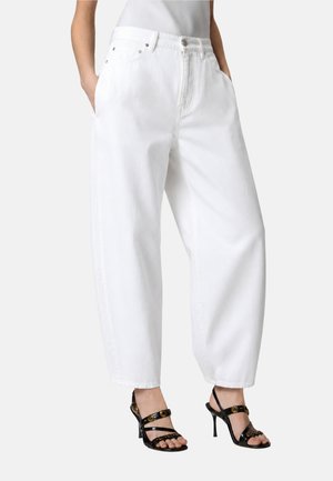 Personne portant un jean blanc ample taille haute avec les mains dans les poches, associé à des sandales noires à talons hauts et à lanières sur un fond neutre.