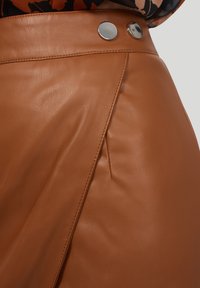 Närbild av en brun läderkjol med sidoflikar och två silverfärgade tryckknappar i midjan, bärs tillsammans med en mönstrad topp.