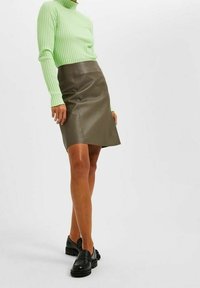 Maglione verde a coste con colletto alto e maniche lunghe, abbinato a una minigonna in pelle marrone attillata e mocassini neri.