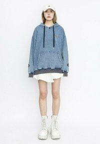 Sudadera con capucha de mezclilla con un bolsillo frontal, cordones negros y texto bordado. Combinada con una falda blanca texturizada y botas blancas gruesas.