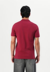 Mand med mørkt hår iført en kortærmet maroon polo-shirt og grå jeans, stående med ryggen til mod en almindelig hvid baggrund.