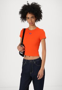 Lyhyt oranssi ribbitekstuuri crop-top, jossa on pyöreä kaulus ja pieni sininen "HUGO" -logo. Yhdistetty tummansiniset korkeat farkut ja musta laukku.