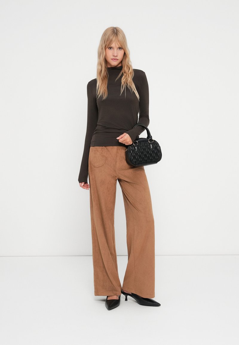 Brązowa bluzka z długim rękawem, beżowe spodnie z szerokimi nogawkami i czarna torebka o fakturze. Obuwie to czarne czółenka z ostrym noskiem. Prosty, współczesny design.