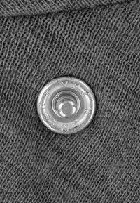 Silberner Metall-Druckknopf auf einem dunkelgrauen Strickstoff, mit einer glatten Textur und eingeprägtem Branding am Rand.