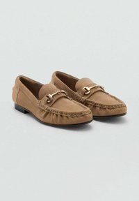 Ett par bruna mockalågsko med detaljer i guldmetall i form av hästbid och sydd design i moc toe, på en neutral grå bakgrund.