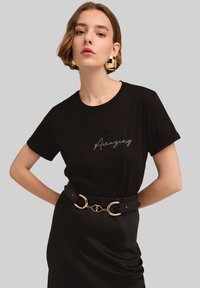 Camiseta de algodón negra con el texto "HAZ QUE HOY SEA Increíble" bordado en contraste. El modelo lleva un cinturón negro con detalles en dorado.