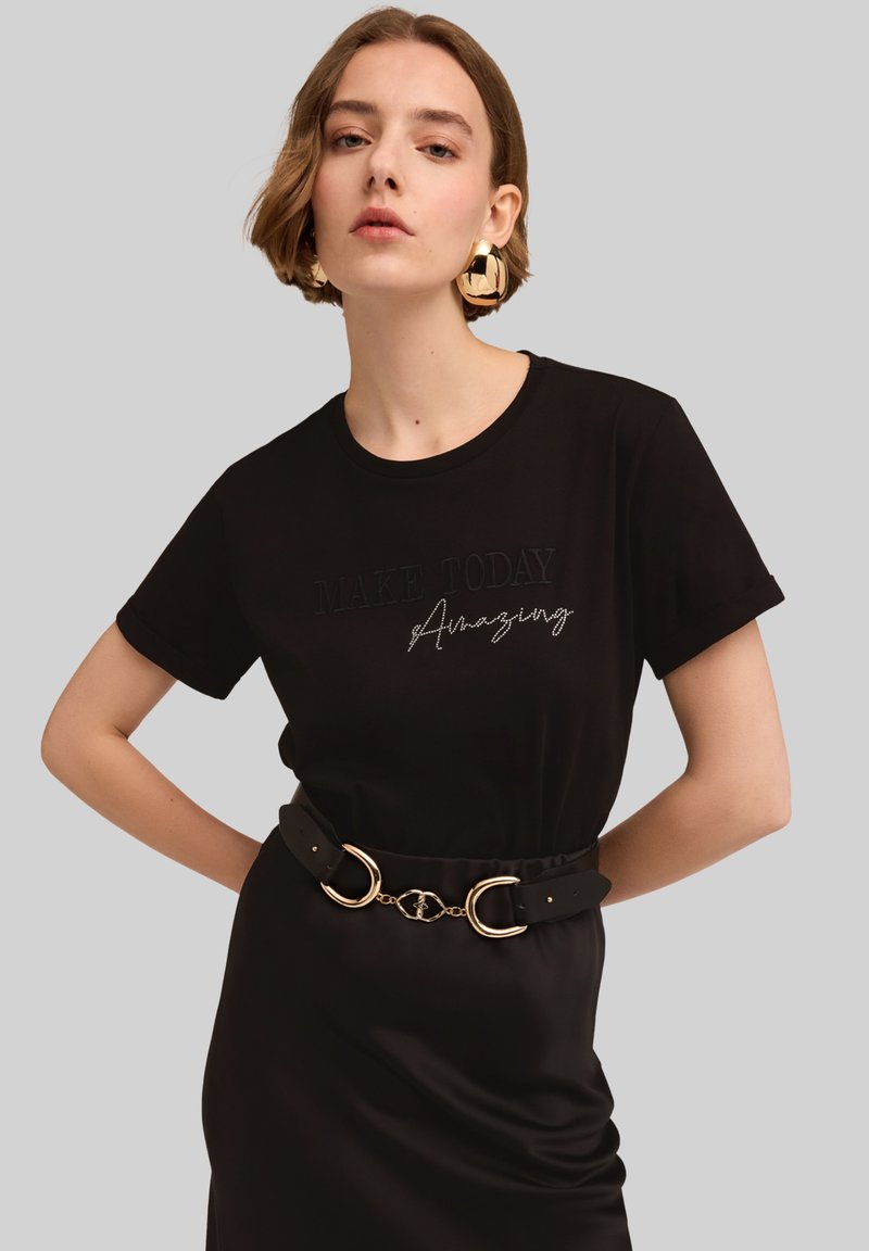 Camiseta de algodón negra con el texto "HAZ QUE HOY SEA Increíble" bordado en contraste. El modelo lleva un cinturón negro con detalles en dorado.