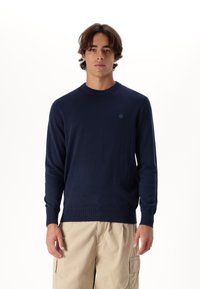 Námořnická modrá pletená mikina s kulatým výstřihem, žebrovanými manžetami a lemem, se malým logem na hrudi. Měkká textura, přiléhavý střih.