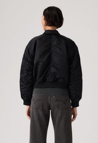 Άτομο που φοράει μαύρο μπουφάν τύπου bomber και σκουρόχρωμο γκρι τζιν, εμφανιζόμενο από πίσω με ουδέτερο φόντο.