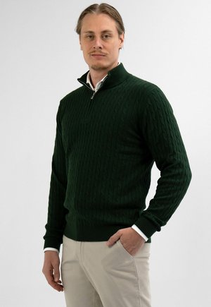 Mann trägt einen dunkelgrünen, grob gestrickten Pullover mit Viertel-Reißverschluss und beige Hosen, steht mit einer Hand in der Tasche vor weißem Hintergrund.