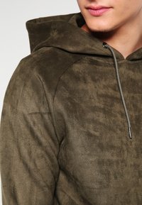 Felpa verde oliva realizzata in morbido tessuto spazzolato, con colletto regolabile e una texture sottile. Inquadratura ravvicinata che mette in evidenza i dettagli del materiale.