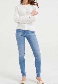 Bílý texturovaný cardigan s kulatým výstřihem a zavíráním na knoflíky, kombinovaný s úzkými džíny světle modré barvy. Ležérní outfit vhodný pro různé příležitosti.