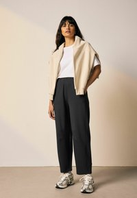 Beżowa krótka kurtka na białej koszulce, w połączeniu z czarnymi spodniami i metalicznymi tenisówkami, stojąca na neutralnym tle.