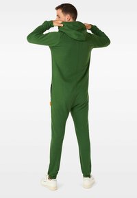 Mono con capucha verde hecho de tela suave, con mangas largas y bolsillos laterales; cuenta con una cremallera y un ajuste relajado. Zapatillas usadas para contraste.