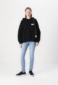 Sweat-shirt à capuche noir avec poche avant et logo blanc, associé à un jean skinny bleu clair et des baskets noires avec une semelle blanche.
