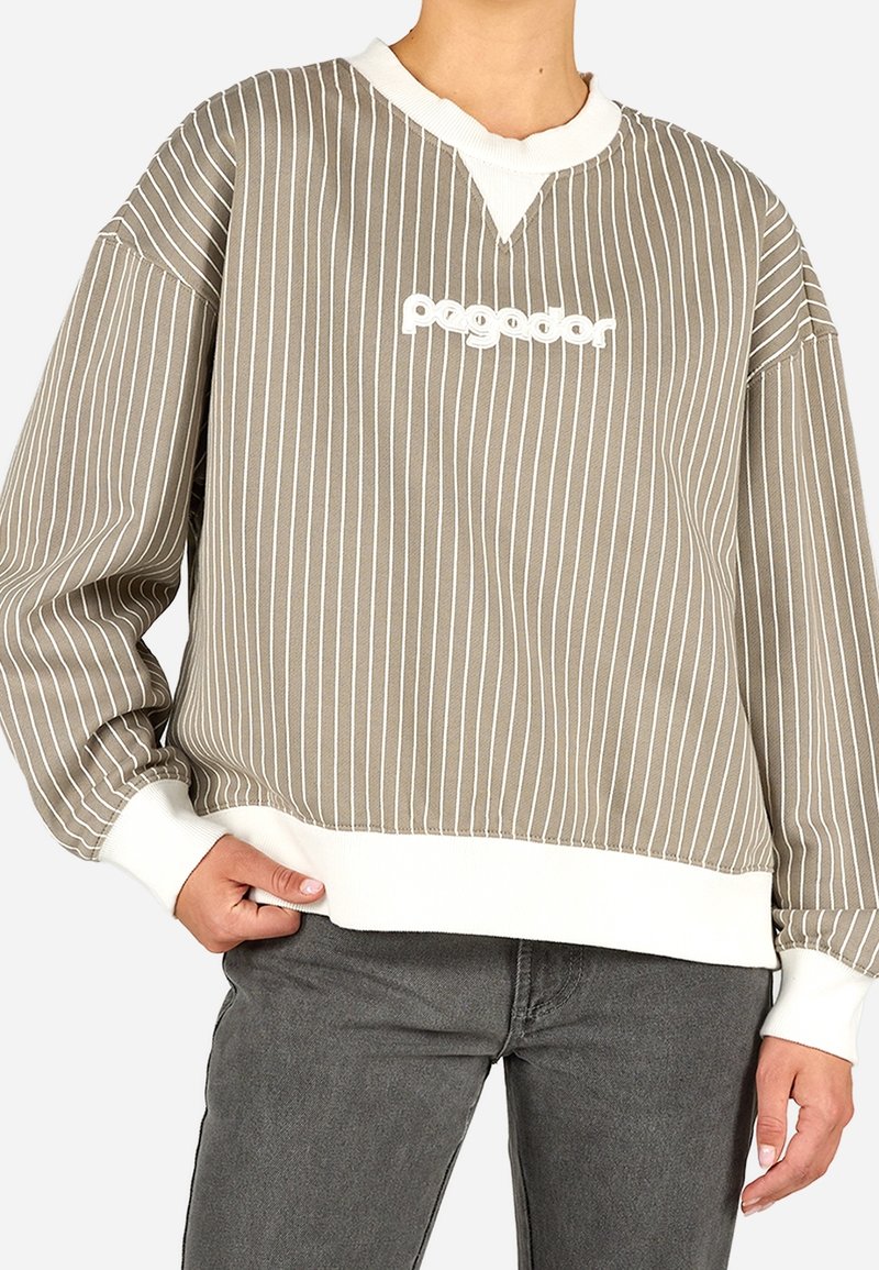 Personne portant un sweat-shirt rayé vertical taupe et blanc avec le texte « pogodr », associé à un jean gris foncé, main posée sur la hanche.