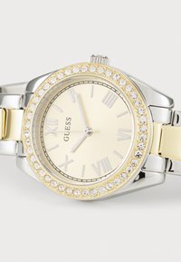Stříbrné a zlaté hodinky s kulatým žlutým ciferníkem, římskými číslicemi a bezel s krystaly. Ocelový pásek.