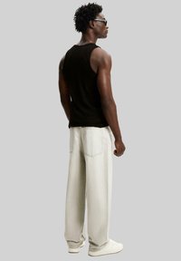 Débardeur noir sans manches, jean gris clair ample, baskets blanches. Le mannequin se tient de côté, affichant une silhouette musclée et des cheveux texturés.