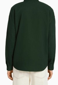 Tmavě zelená texturovaná dlouhý rukáv košile s límečkem, detaily na zadním sedle a manžetami na knoflíky, v kombinaci se světlými kalhotami.