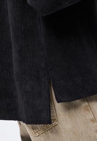 Krupni plan crnog rebrastog topa s bočnim prorezom nošenog preko bež traperica s vidljivim šavovima na stražnjim džepovima.