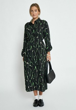 Zwarte jurk met een groen abstract patroon, met knoopsluiting bij de kraag, lange mouwen, een geaccentueerde taille en een rechte snit, gecombineerd met een zwarte tas en loafers.