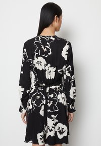 Schwarzes Kleid mit großen weißen Blumenmustern, langen Ärmeln und einer gerafften Taille, die mit einem passenden Band am Rücken befestigt ist.