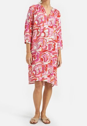 Femme portant une robe à motifs floraux rose, rouge et blanc, longueur genou, manches 3/4, et des sandales beiges ouvertes à l’avant, debout sur fond blanc.