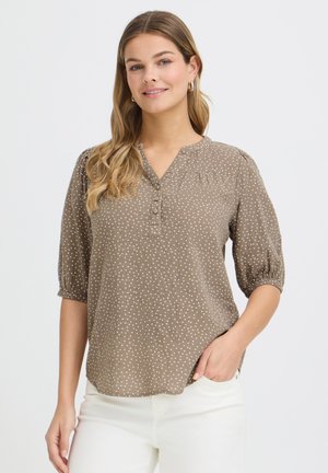 Žena s dlhými blond vlasmi, ktorá má na sebe taupe blúzku s bodkami a trojštvrťovými rukávmi a biele nohavice, usmieva sa s jednou rukou v vrecku.