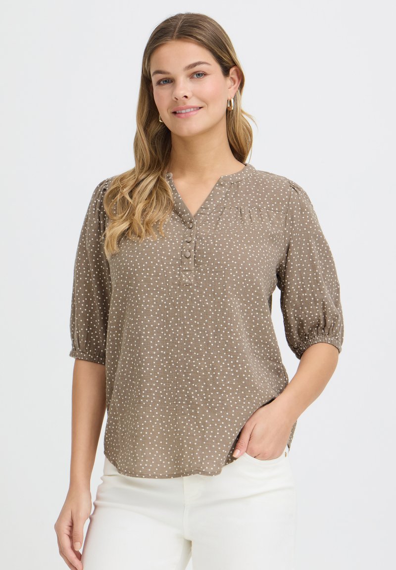 Femme aux longs cheveux blonds portant un chemisier taupe à pois avec des manches trois-quarts et un pantalon blanc, souriante avec une main dans la poche.