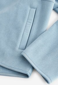 Svijetloplava flis jakna od mekog, strukturiranog materijala, s glatkim džepom i donjim rubom, šivenim rubovima i produženim rukavima.