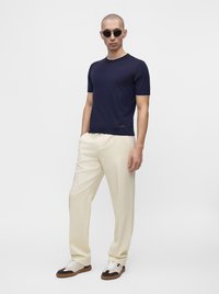 Homme portant une chemise bleu foncé à manches courtes, un pantalon crème, des lunettes de soleil et des baskets bicolores, debout les mains dans les poches sur un fond uni.