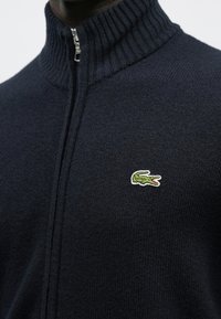 Camisola azul-marinho com fecho de correr e gola canelada, com um logotipo de crocodilo verde no lado esquerdo do peito. Feita de tecido de malha texturada.