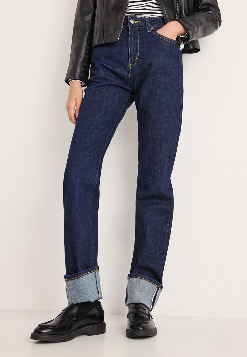 Tumesinised teksad sirge jala lõikega, millel on kergemast teksast ülespööritud mansetid ja kollane õmblus taskutel ning õmblustel.