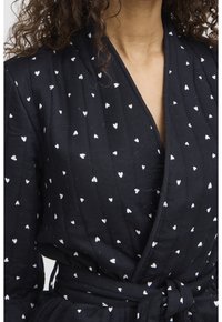 Robe noire enroulée avec des motifs de cœurs blancs, en tissu texturé. Présente une taille à nouer, un décolleté en V et des plis verticaux.