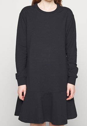 Žena v dlouhých černých šatech s kulatým výstřihem a volánkovým lemem, stojící před jednobarevným světlým pozadím.