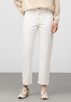 Person, die weiße Jeans mit hohem Bund und geradem Bein, einen beigen Pullover sowie beige-weiße Schnürsneakers trägt und auf einem grauen Boden steht.