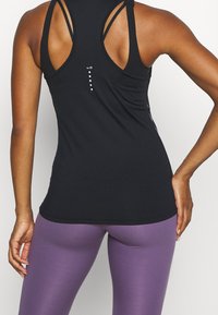 Débardeur de sport noir avec plusieurs bretelles dans le dos, présentant une finition texturée et un logo discret sur le côté. Associé à un legging violet.