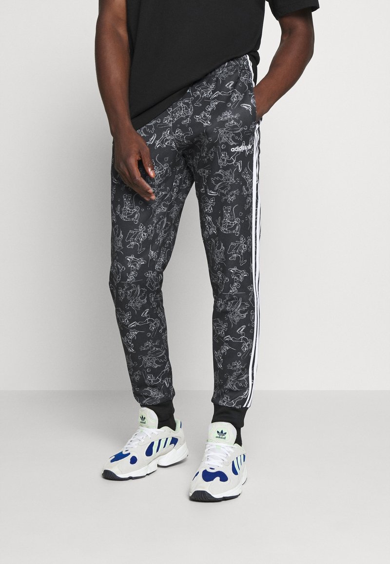 Man som bär svarta Adidas-joggingbyxor med vita linjetecknade figurer, vita Adidas-sneakers och en svart t-shirt mot en neutral bakgrund.