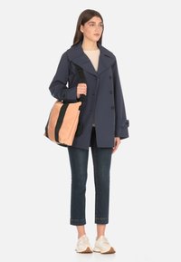 Impermeabile blu navy con un ampio colletto, bottoni doppi e maniche larghe; porta una borsa a tracolla rosa e nera; jeans scuri; scarpe chiare.