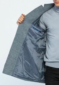 Cappotto in lana grigio chiaro con una fodera interna grigia liscia. Presenta una tasca nascosta con un bordo decorato. Silhouette sartoriale e apertura frontale.