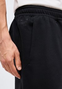Κοντινή λήψη του χεριού ενός ατόμου που ακουμπά δίπλα σε μαύρο ελαστικό παντελόνι jogging με λεπτό ανάγλυφο κείμενο κοντά στην τσέπη.