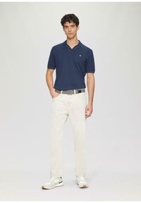 Marineblaues Poloshirt mit V-Ausschnitt, cremefarbene Jeans und weiße Sneaker mit grauen Akzenten. Ein einfacher Gürtel rundet das Outfit ab.