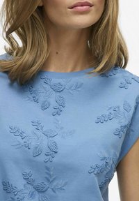 Jasnoniebieski bawełniany top z fakturową haftowaną florą w ciemniejszym odcieniu niebieskiego, okrągłym dekoltem i krótkimi rękawami.