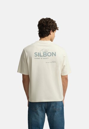 Hombre con cabello rizado que lleva una camiseta beige de manga corta con el texto "The Silbon Classic Society" en la espalda, combinada con vaqueros azules.