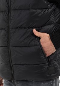 Schwarze Steppweste mit diagonal gesteppten Abschnitten, einem Reißverschluss vorne und einer Seitenasche mit einem kleinen Logobus am Saum. Strukturiertes Material.