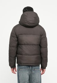 Person, der bærer en sort pufferjakke med hætte og blå jeans, vender væk mod en ensfarvet hvid baggrund.