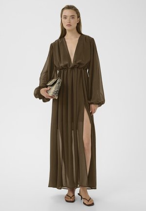 Vrouw draagt een lange, bruine, doorschijnende jurk met diepe V-hals, hoge split, pofmouwen, houdt een clutch van slangenleer vast en draagt zwarte sandalen met bandjes.